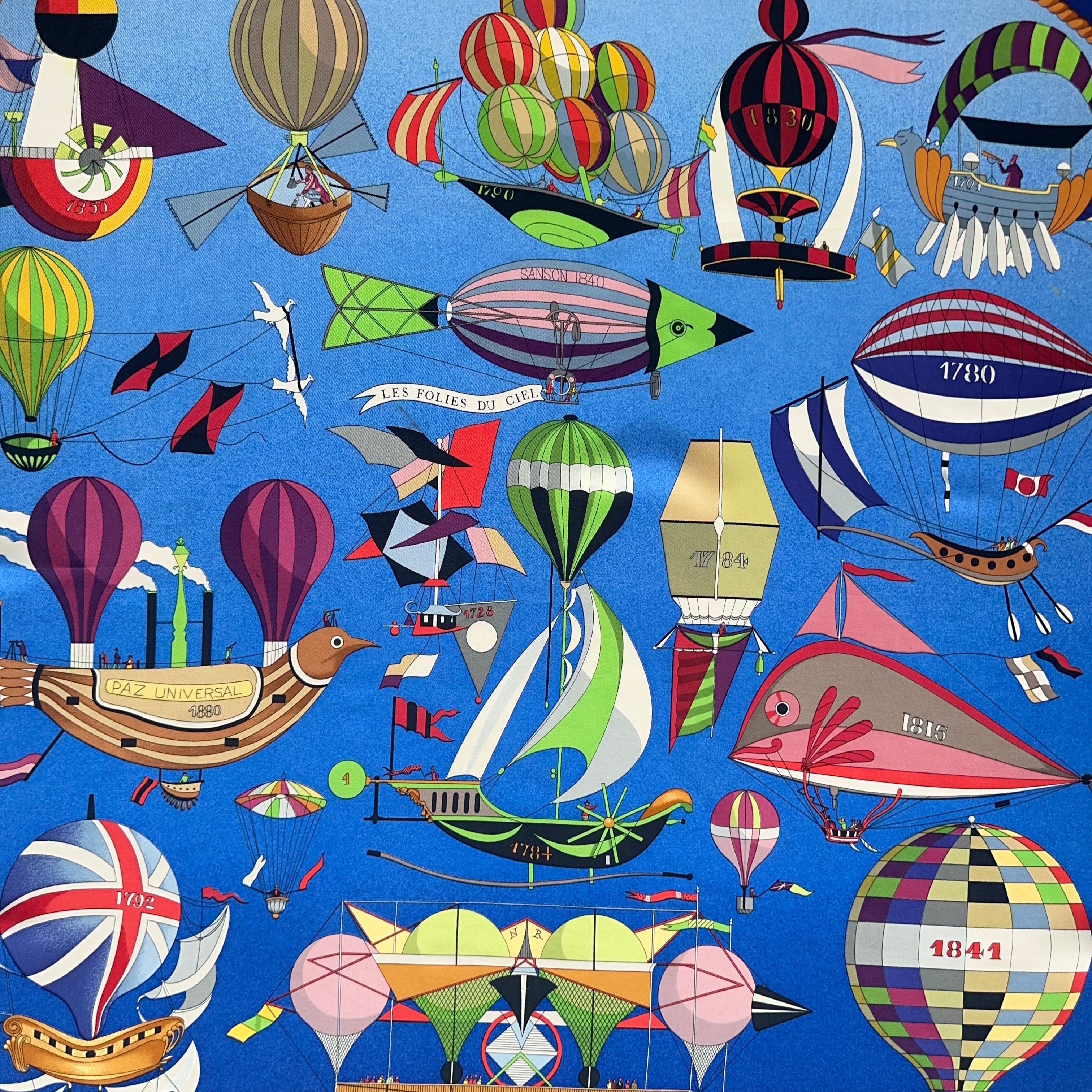 Zoom sur la partie supérieure du foulard : ballons à hélice, montgolfières rayées et machines volantes fantaisistes rouges et vertes sur fond bleu.